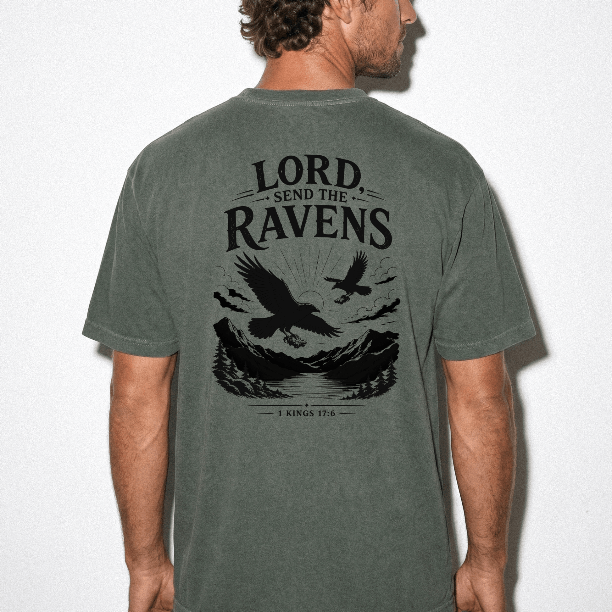 Back view of a man wearing the Lord, Send the Ravens Christian graphic tee featuring ravens, mountains, and the Bible verse 1 Kings 17:6 on a vintage olive green unisex shirt.