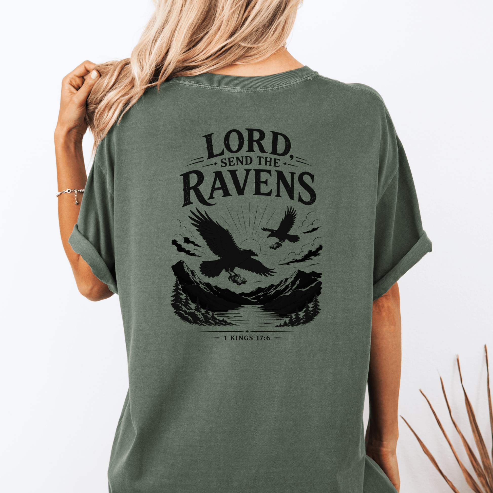 Woman wearing the Lord, Send the Ravens Christian graphic tee featuring ravens, mountains, and the Bible verse 1 Kings 17:6 on the back of a unisex faith-inspired shirt.